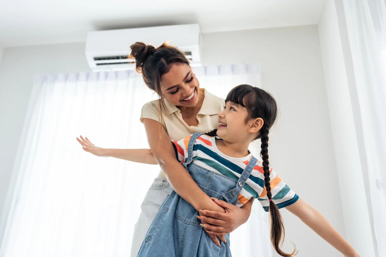 A woman and her young daughter engage with a ductless air conditioner, showcasing a playful moment in their living space.